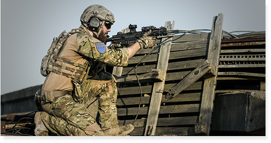 A soldier in camouflage and gear is aiming his rifle.